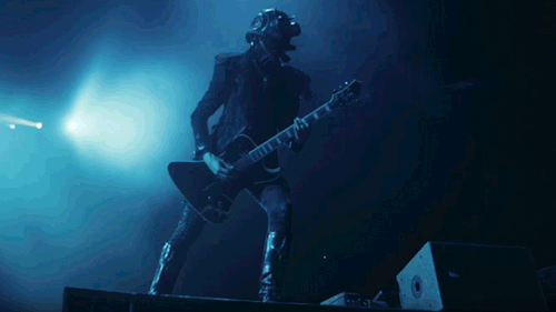 Phantom Ghoul plays guitar on the edge of the stage, leaning forward toward the crowd. He snaps his head from side to side with the beat. 