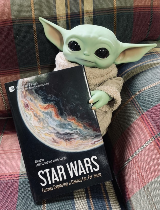Grogu sitting on a plaid chair holding a copy of the book Star Wars: Essays Exploring a Galaxy Far, Far Away (Vernon Press, 2023), edited by Emily Strand and Amy H. Sturgis, with cover art (a watercolor painting of a planet) by Emily Austin.