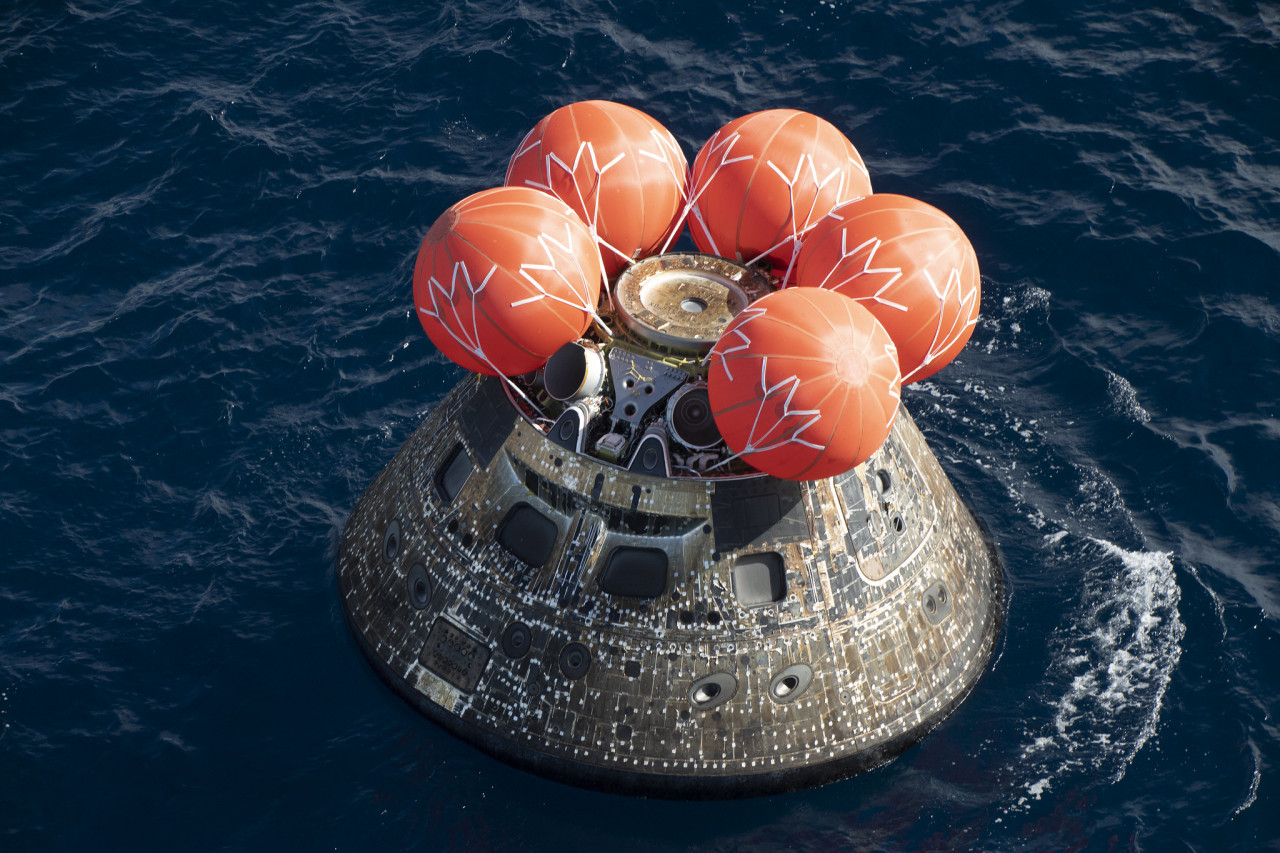 The Orion spacecraft floats in the ocean shortly after splashdown. Five orange airbags are strapped to the top of the capsule. The outside of the spacecraft appears silver and brown. The ocean is a deep blue. Credit: NASA