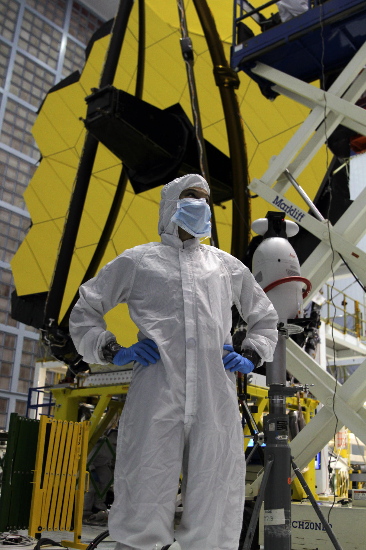 Sophia stands with her hands on her hips in front of Webb’s golden mirrors. She is dressed in a head-to-toe white jumpsuit, blue latex gloves, and a disposable face mask. Various construction equipment is scattered around her. The background is a large, open clean room with a ventilation system on the back wall, made up of panels of small light-pink squares. Credit: NASA