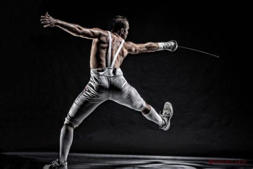 modernfencing - [ID - a shirtless epee fencer lunging away from...