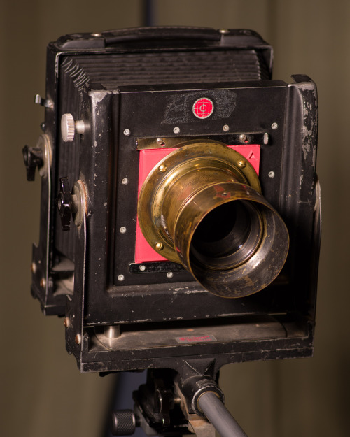 An old 4x5 camera has a large brass Petzval lens mounted to the front. The lens board is thicker than normal to allow the brass flange to overhange the edges of the lens board mounting area.
