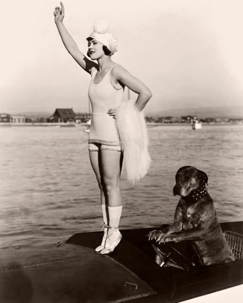 Gloria Swanson (1899 - 1983) stands on tiptoes on the prow of a motorboat while Teddy the dog sits with his paws on the steering wheel in a still from director Clarence G Badger’s film Teddy at the Throttle, 1916.