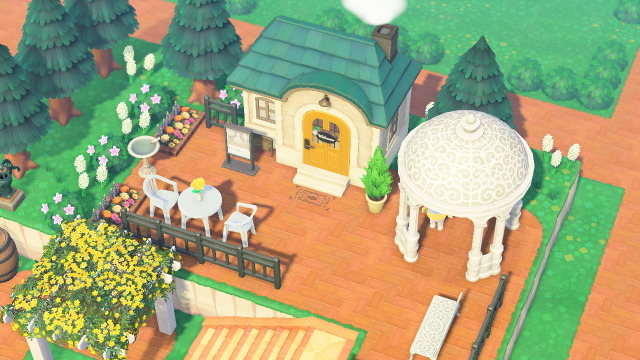 A side-profile of the exterior of the chalet. a white-wood pergola with yellow flowers gives rise to a brick staircase leading to the storefront. On this level, there is an angelic white gazebo and iron garden bench painted white. 