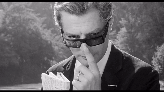 A scene from 8½. Guido Anselmi played by Marcello Mastroianni taps his nose while holding a folded newspaper against his suit and peering over his sunglasses