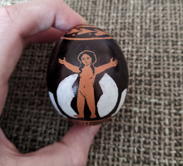 An egg that has been drawn on to resemble an ancient Greek red figure vase depicting Helen, small but fully formed, standing with her arms raised between two halves of an egg shell. This is from a central angle.