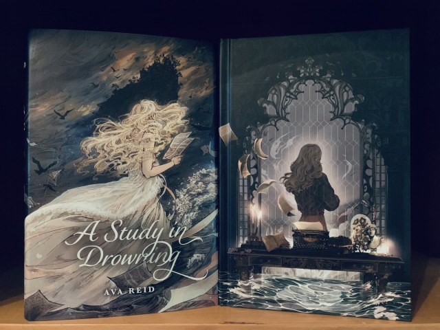 Shown is the Owlcrate special edition of A Study in Drowning. The paper cover has artwork depicting a young woman with a book in the wind, with books and ravens swirling around her, water, and a menacing building in shadow behind her. The naked hardback has artwork depicting a woman at an ornate window, standing behind a desk with an antique bust, typewriter, and books on it, illuminated by candles. Water is rising in the foreground. Photo by AHS.
