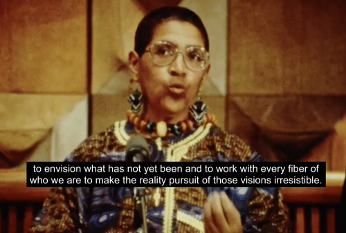 Audre Lorde stands at a podium accepting her award and appointment as State Poet of New York for 1991-1993. She is dressed formally in a colorful tunic. She raises one hand in front of her for emphasis and concludes, "to envision what has not yet been and to work with every fiber of who we are to make the reality pursuit of those visions irresistible."