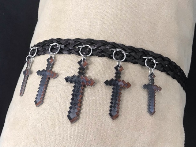 photo of a woven leather choker embellished with five minecraft netherite sword charms dangling from the front pointing down. The swords are arranged bu size with the largest in the center and the smallest towards the outside.