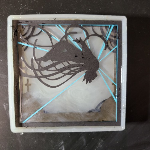 More layers of paper set in the resin. The silhouette of the back flip is the top-most layer. Below can be seen a layer of rooftops and thin blue lines. The resin has thin bits of white ink swirled into it.