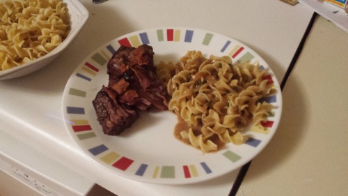 A plate of pot roast, topped with bacon, along with a side of egg noodles and brown sauce.