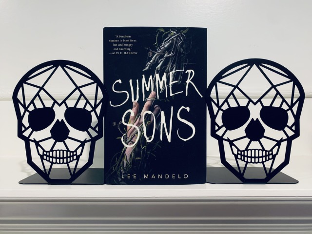 The picture shows SUMMER SONS (with cover art depicting a human hand and skeleton hand reaching out for each other) on a white mantel, bracketed by black skull bookends. Photo by AHS.