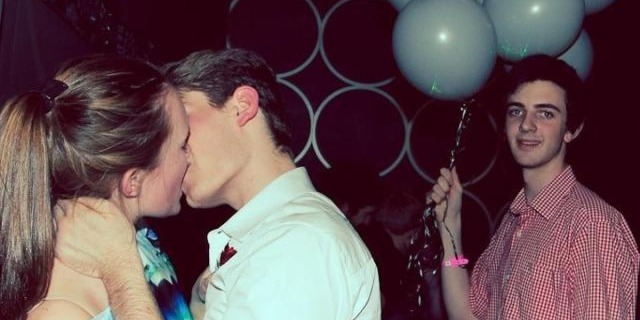 two people kissing, while a man stands behind them holding a balloon, smiling uncomfortably