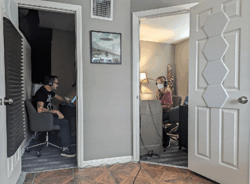 A photo of Wil Wheaton and Gabrielle De Cuir in separate studios at Skyboat Media. Wheaton is animated and looks up from the mic to shrug.