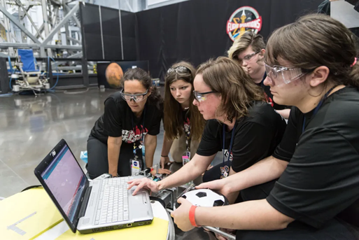 In the right foreground, five people huddle around a laptop computer wearing clear protective goggles and black t-shirts. A tall, black divider with a flight operations insignia stands in the background next to a large machine. Credit: NASA