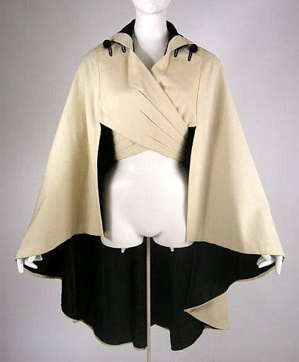 a wrap cape on a dress form. it's about hip-length, flared, longer in the back, and collared. it features 2 lengths of fabric that cross over the torso and close in the back.