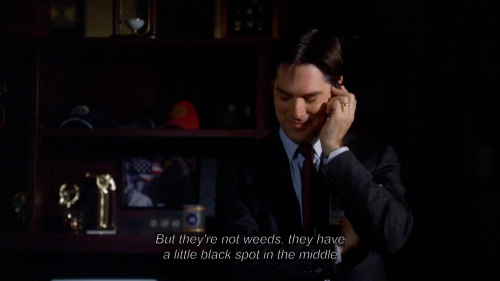 In a decorated office, a man in a suit has one arm wrapped around his midsection and the other holding a phone to his ear. He is looking down and smiling, mid-laugh. The subtitle at the bottom of the image, which is in italics, reads: "But they're not weeds, they have a little black spot in the middle"