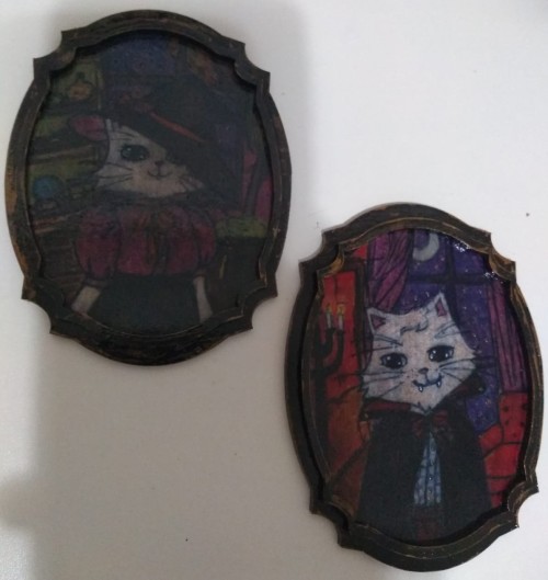 Two tiny frames, one with a drawing of a cat dressed as a witch and the other with a drawing of a cat dressed as a vampire. Both are colored and humanoid.