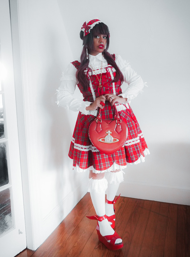 black person red hair wearing a plaid lolita dress, white blouse, lace knee high socks, red sandals with wings and red vivienne westwood heart purse