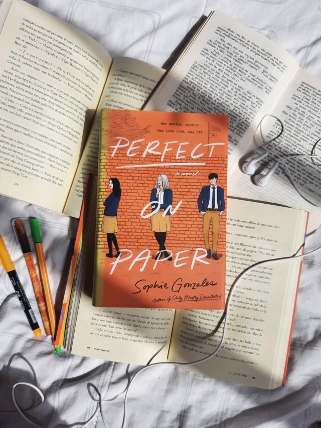 a copy of Perfect On Paper by Sophie Gonzalez over three open books with earphones wires. some pencils orange pencils are in left side of the book. white blanket as background. 