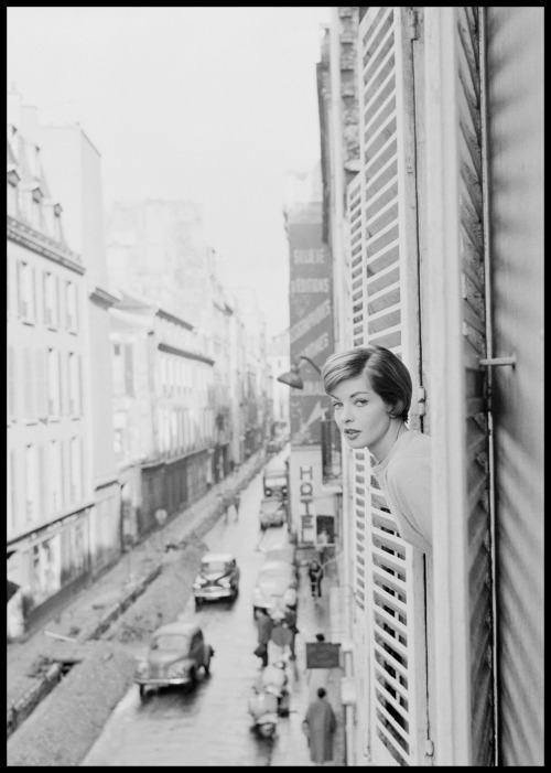 A model looks out of the window of her Paris hotel, overlooking a busy street below; 1959