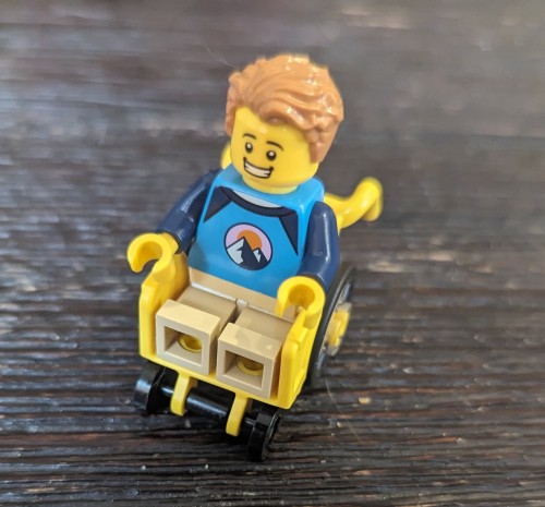 [First Image description: a close-up photo of a  Lego™ figure in a plastic molded Lego wheelchair. end description]