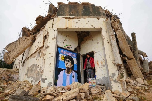 Artist Aziz Asmar stands by his painting of Diego Maradona on the wall of a destroyed home. Binnish,