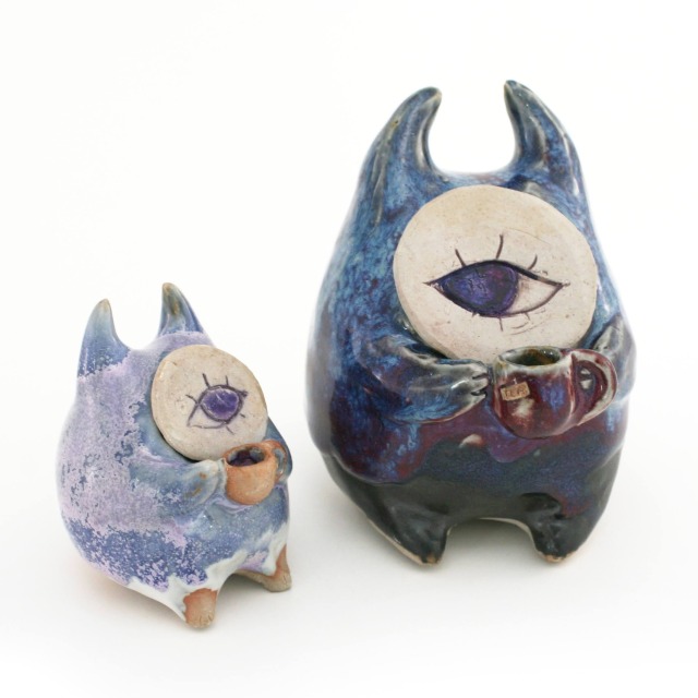 Two ceramic sculptures of small round horned creatures, sitting up and holding mugs in their paws, peering at each other with single eyes in round unglazed faces. One is smaller and glazed in lavender and white, while the larger is glazed in blue, magenta and black.