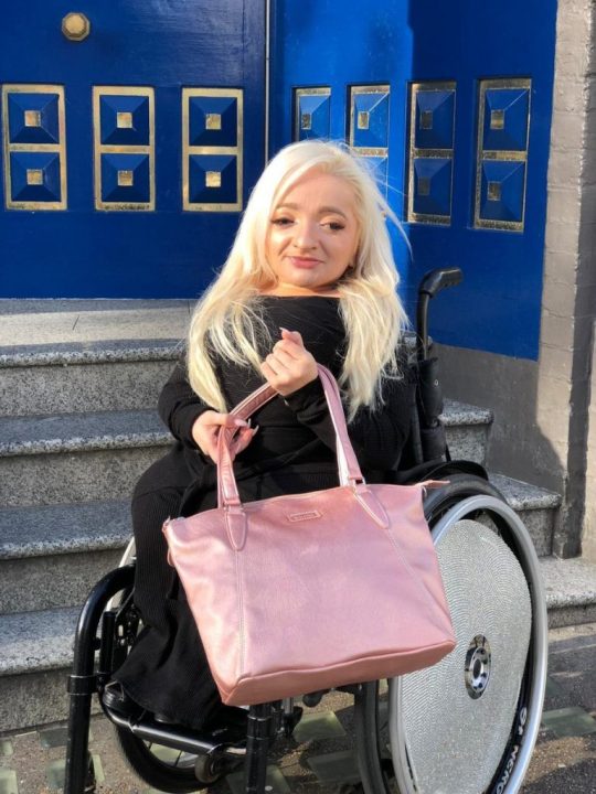 Photograph of Sam Renke holding a bag from her Samantha Range in a Rose Gold colour, as she sits in her wheelchair at the base of a set of granite stairs