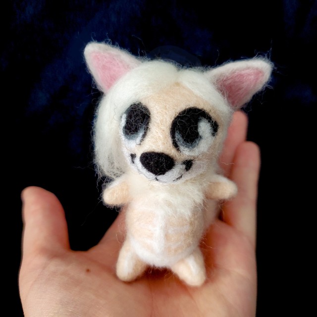 A photo of a needle felted sculpture of a fox-like creature named Menoes. She is peach colored with long, silver hair and white markings that look like bones. She is being held by a hand.