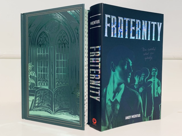 Shown is the Unplugged Book Box special edition of FRATERNITY. The naked hardback has a foiled pattern of an open tome underneath a Gothic window. The book cover has artwork depicting four young men who are classmates. Both have green and black colors. Photo by AHS.