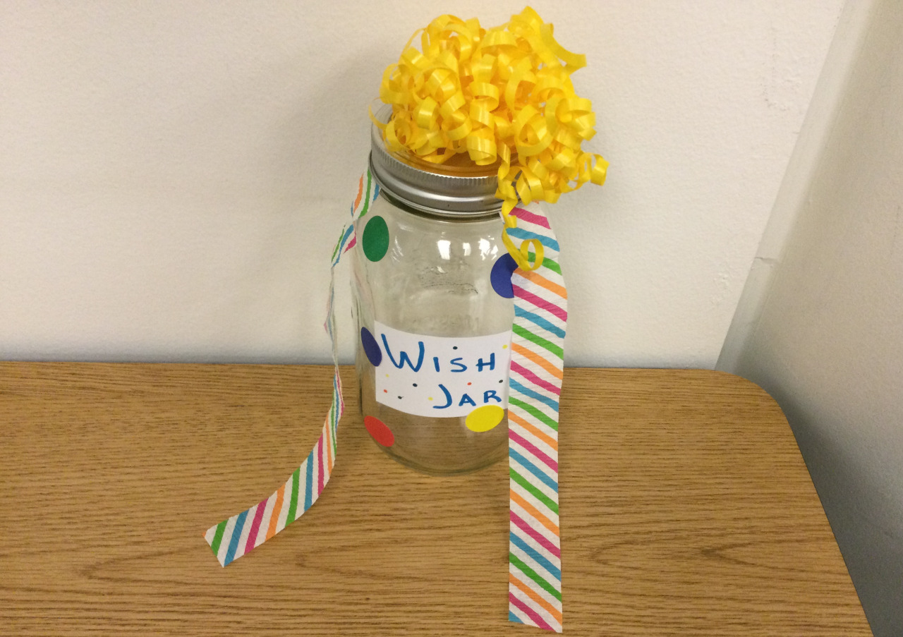 A mason jar decorated with colorful circle stickers, yellow curling ribbon, and crepe paper. It is labeled wish jar