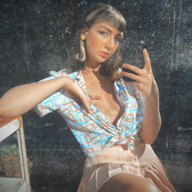 Peaches, a pale thin femme, posing in a mirror with her left hand resting on her chest, wearing a white button up shirt with pastel shapes on it un buttoned to reveal cleavage