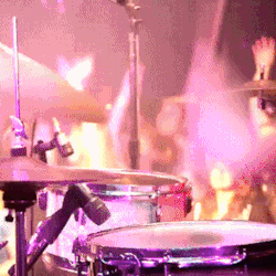 a person on stage playing the drums, with the crowd visible behind the drumset, dancing and waving their hands. multicolored lights flash, lighting up the scene