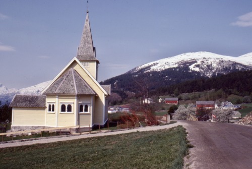Bybildet om våren med kirke, røde fjøs og snødekte fjell, Liabygda, Møre og Romsdal, Norge, 1981.
