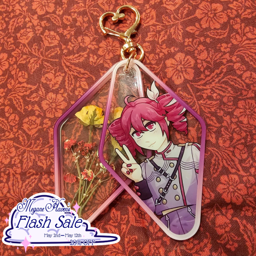A two piece acrylic charm shaped in a long diamond shape. The front piece has an illustration of Kasane Teto (SynthV design) with a cat-like smile and peace sign hand gesture. The back piece holds small dried roses inside.