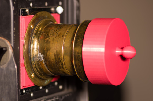 Large format brass lens from 1850 mounted to a red 3d-printed lens board with a red 3d-printed lens cap over the hood.