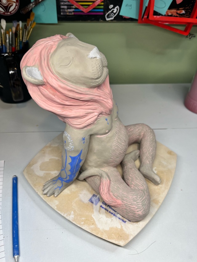 a clay sculpture of a humanoid figure with the head and tail of a lion. the mane and body hair are pink and he has blue tattoos. he\'s got pink top surgery scars