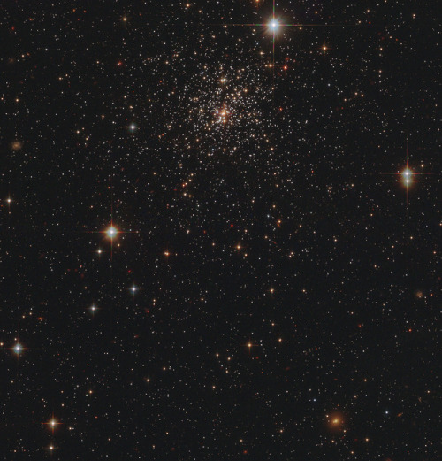A star cluster glowing softly in the void with faint, reddish background galaxies in the spaces betw