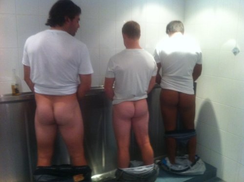 Three men pissing in a urinal trough with their pants down to their ankles.  One checking out his bu