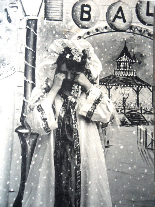 Lovely postcard of woman in her Edwardian coat with hood, who stands at the entrance of a “Ball Masq