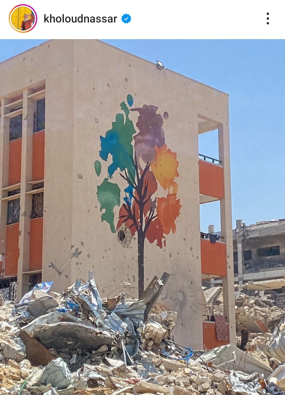 A colorful tree mural on the side of a damaged building. All the surrounding buildings are reduced to rubble.