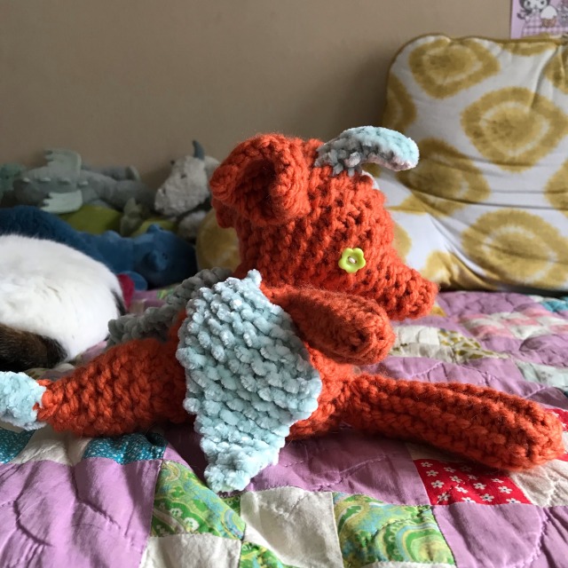 An orange and blue knitted dragon with flower button eyes and a pointy tail sits on a purple quilt 