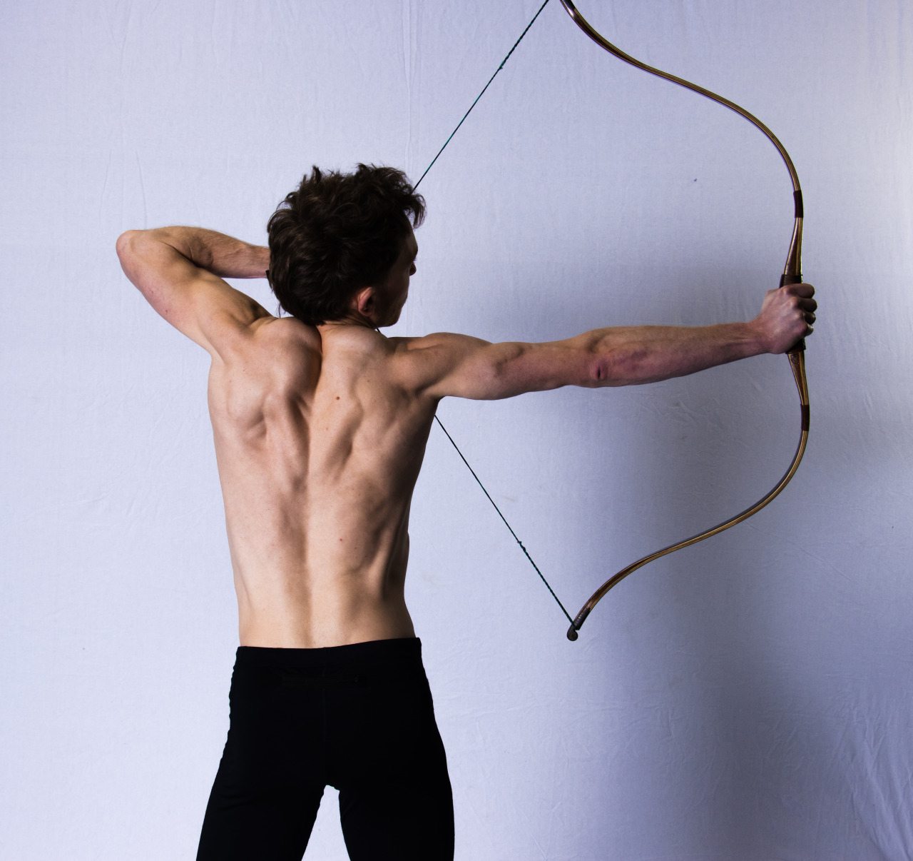 a photograph of a shirtless man drawing a 120lb bow, showing muscle engagement.