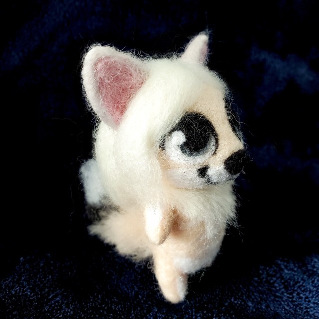 A photo of a needle felted sculpture of a fox-like creature named Menoes. She is peach colored with long, silver hair and white markings that look like bones. She is against a dark blue background.