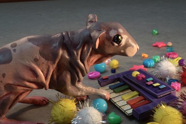 A photo of a hand-sized painted clay figurine of Neil the hairless rat, playing a small purple synthesizer. The photo is taken slightly from above, showing Neil\'s left side.  The knobs, wheels and black keys of the synth are in the colors of the rainbow. The figurine has its right forepaw raised, resting on the keys.  The figurine sits on a grey vinyl floor, and is surrounded by colorful glittery pompoms and plastic beads in a variety of shapes laid out around it. Some of the beads are shaped like stars and musical notes.