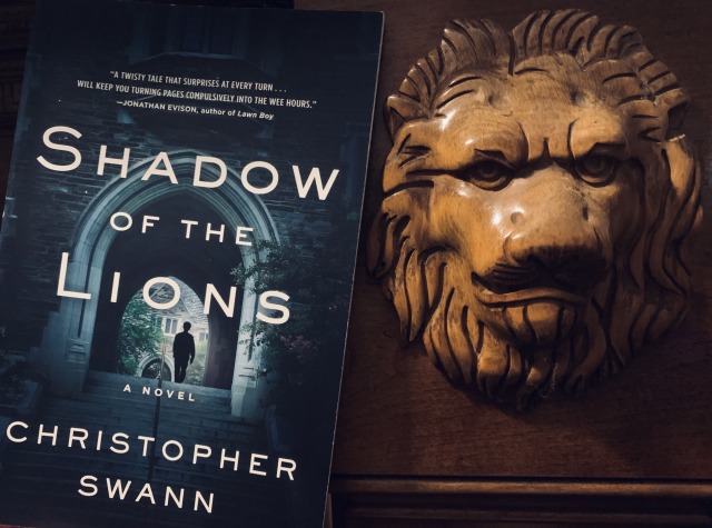 Pictured is SHADOW OF THE LIONS (with cover art depicting a lone figure walking under an arch on a campus) next to a lion carved in wood. Photo by AHS.