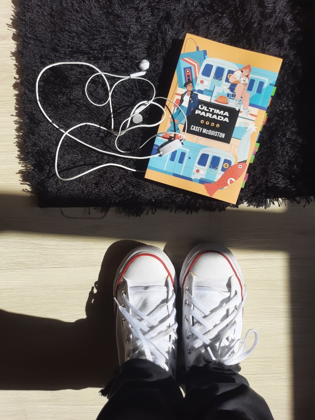 a copy of One last stop by Casey McQuiston on a fluffy black rug with earphones next to it. in the bottom part of the photo, the tip of white converse all-star.