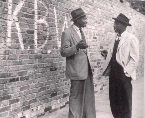 spiritsdancinginthenight:  Two Jamaican men talking to one another, while behind a wall has written 