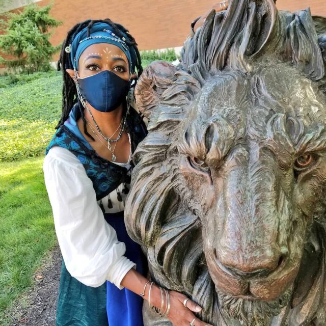 Seneca, dressed as an elven wizard wearing vivid shades of blue and teal, looks into the near distance as they embrace the head of a life-sized bronze lion statue.